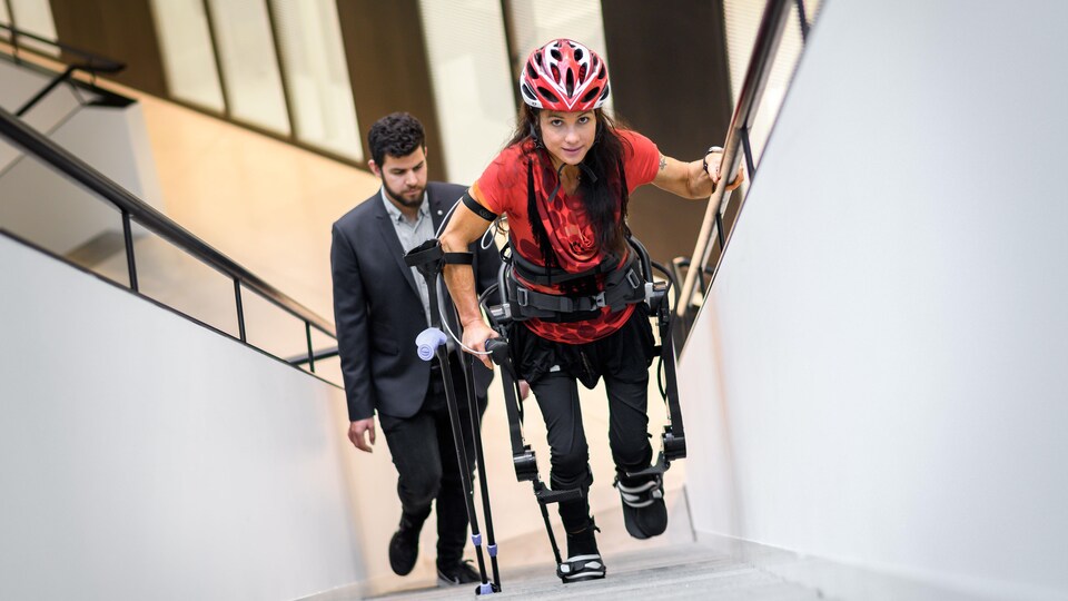 L'athlète monte des marches avec un casque, un gilet rouge et un exosquelette.