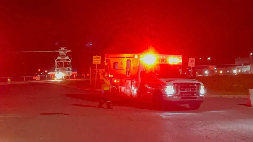 Une photo très rouge prise le soir. On voit une ambulance les feux allumés et derrière un hélicoptère qui se pose ou décolle de la piste d'un héliport.
