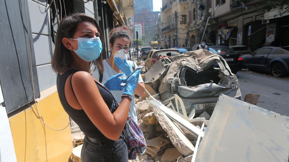 De jeunes Libanaises portant un masque et des gants dans une rue dévastée par les explosions.