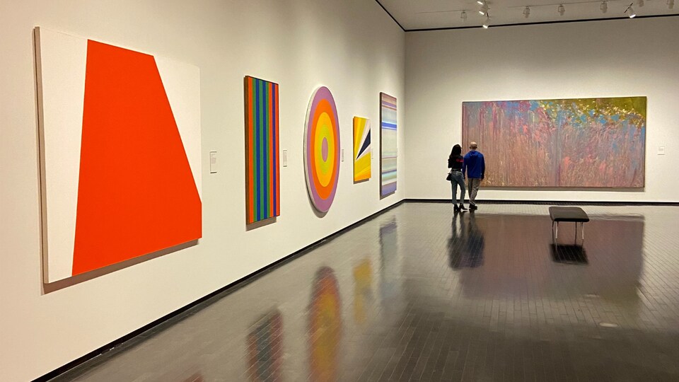Deux visiteurs qui marchent dans une pièce où des peintures colorées sont accrochées aux murs.