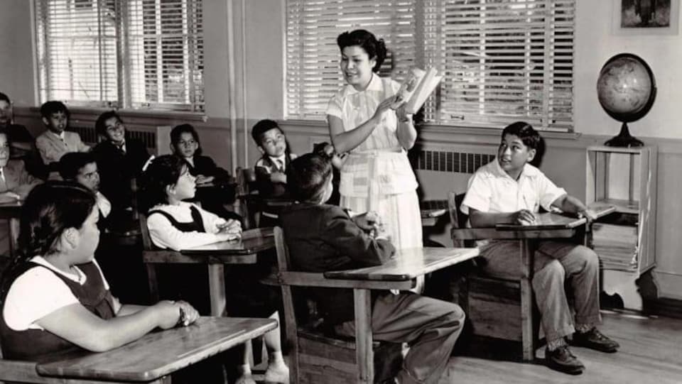 Une femme tient un livre debout au milieu d'une classe composée d'élèves autochtones.
