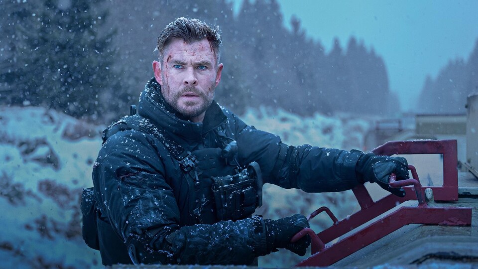 Un homme s'accroche sur le côté d'un train, dans un paysage enneigé, dans une image d'un film d'action. 