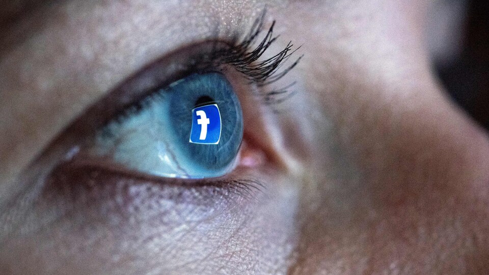 Une femme se tient devant un écran d'ordinateur qui affiche le logo du réseau social Facebook. Ce logo est reflété dans son œil.