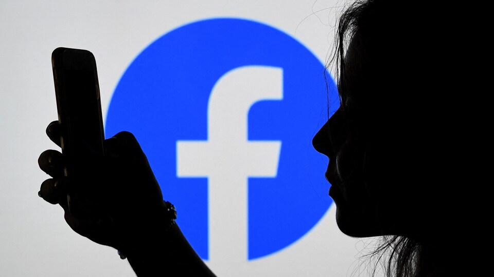Une jeune fille regarde un écran de téléphone intelligent devant un logo géant de Facebook.
