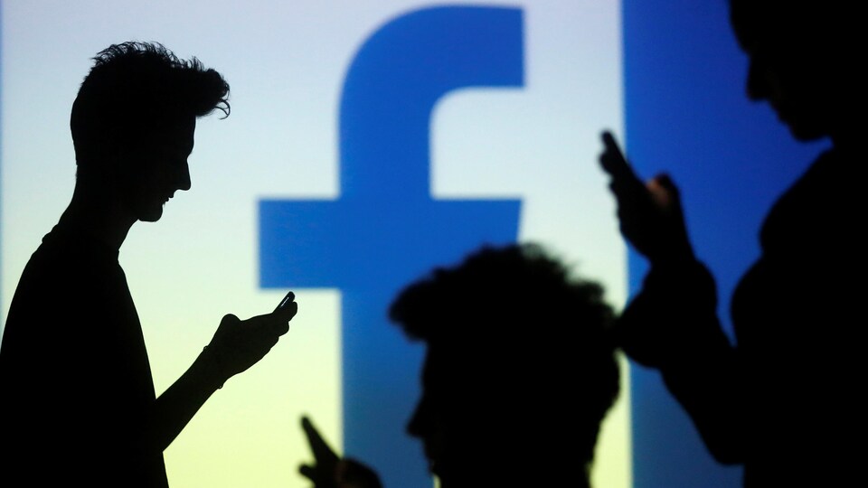 On aperçoit des silhouettes de personnes tenant des téléphones intelligents devant la projection grand format du logo de Facebook.