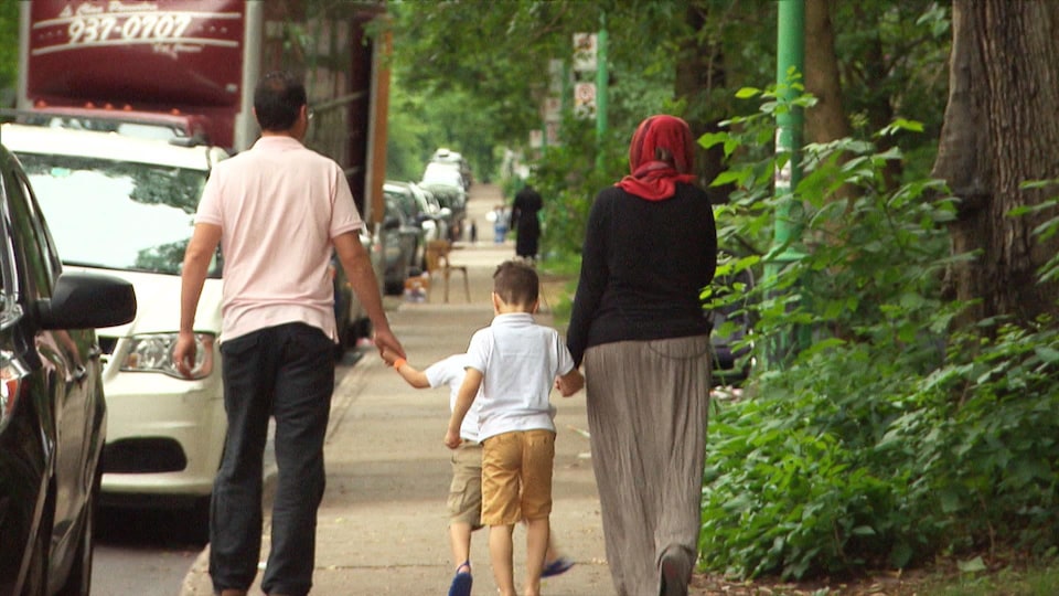 Un homme marchant à gauche sur le trottoir et portant un t-shirt beige tient la main d'un enfant, qui marche au centre du trottoir. Une femme portant un hijab rouge marche à droite sur le trottoir et tient la main d'un second enfant.