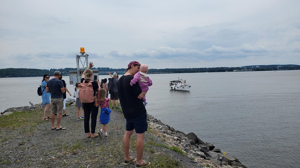 Un dizaine de personnes près de la rive qui attendent la famille Savard-Legault. Au loin se trouve le voilier des Savard-Legault. 