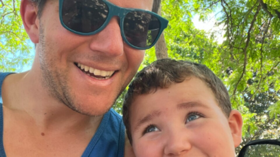 Vincent Hachey porte des lunettes de soleil et pose pour un selfie, avec son fils qui porte un t-shirt de Batman. Le petit garçon pose sa tête contre l'épaule de son père. En arrière-plan, les feuilles des arbres font écran au soleil.