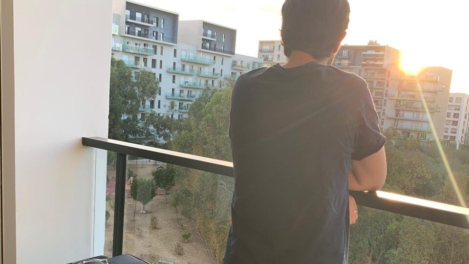Un homme, installé sur un balcon avec ses valises, regarde le soleil qui se couche derrière les tours d'habitation.