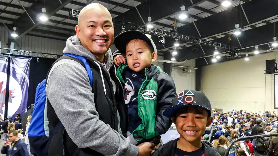 Un homme avec un enfant dans les bras. Un autre enfant, un peu plus vieux, se trouve à ses côtés. Ils portent fièrement les couleurs des Jets de Winnipeg.