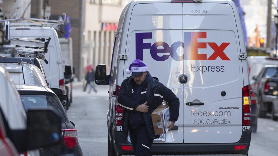 Un livreur porte une tuque de Noël et transporte un colis près d'un camion de Fed Ex.