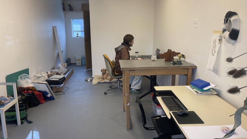 Un jeune homme, flanqué de son chien, est assis devant un ordinateur dans son studio d'artistes.