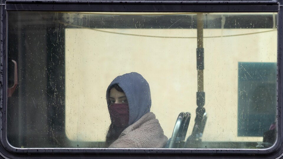 Une femme qui porte un masque est assise dans un autobus et regarde le photographe à travers la fenêtre.