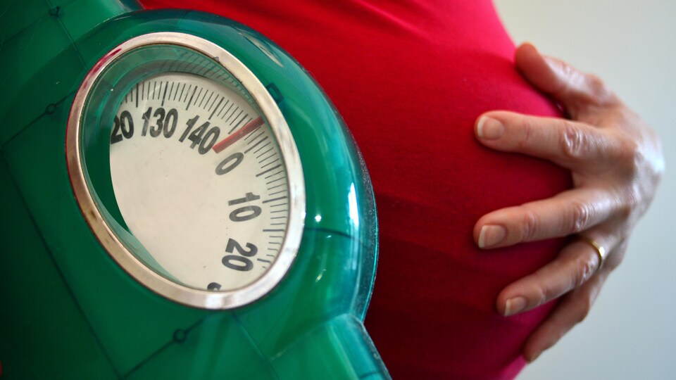 Une femme enceinte tient un pèse-personne et place son autre main sur son ventre.