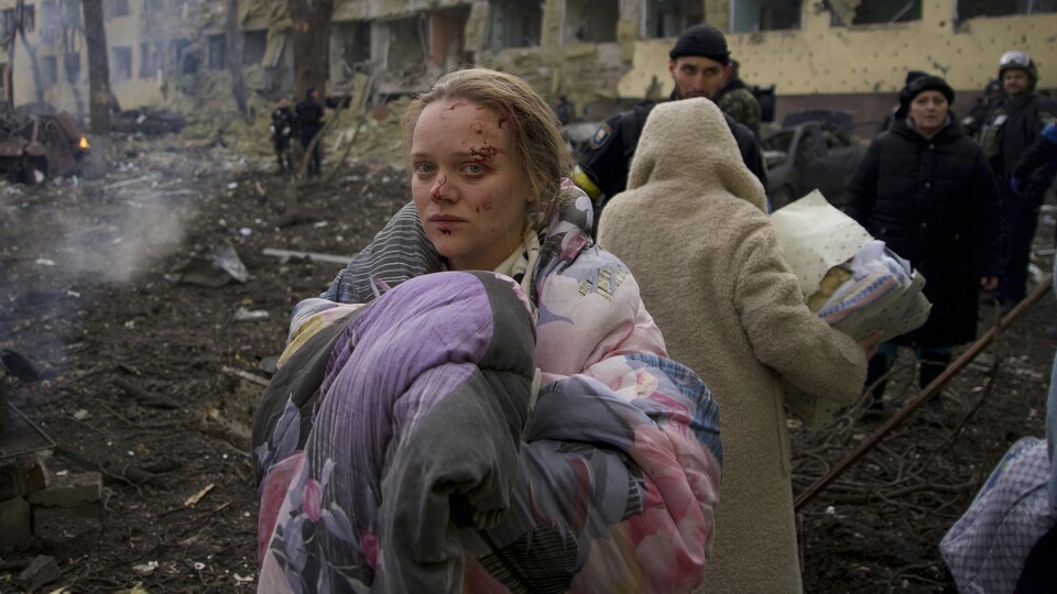 A woman with a facial injury moves outside near a damaged building. She is covered with a blanket.