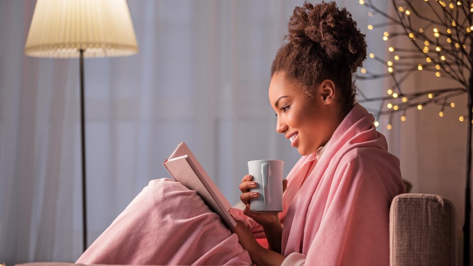 Une jeune femme lit un livre, assise dans un sofa avec une tasse dans la main.