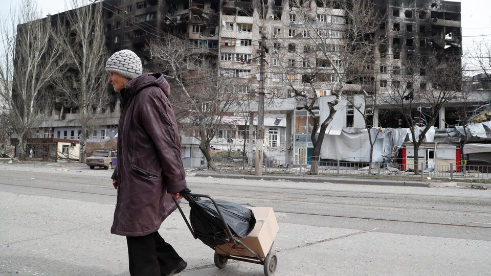 Une femme âgée marche avec un chariot devant un grand immeuble à logement endommagé par les bombes. 