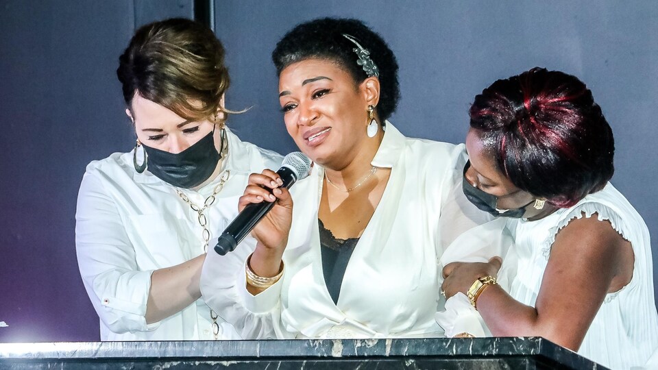 La femme de Paul Mukendi tient un micro dans les mains et est épaulée par deux femmes.