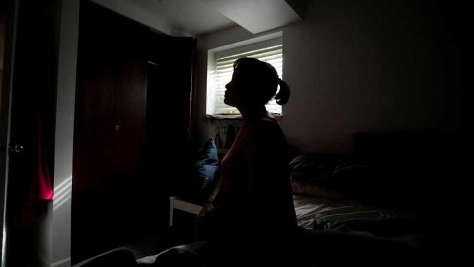 Une femme est assise sur son lit, dans la pénombre du chambre à coucher.