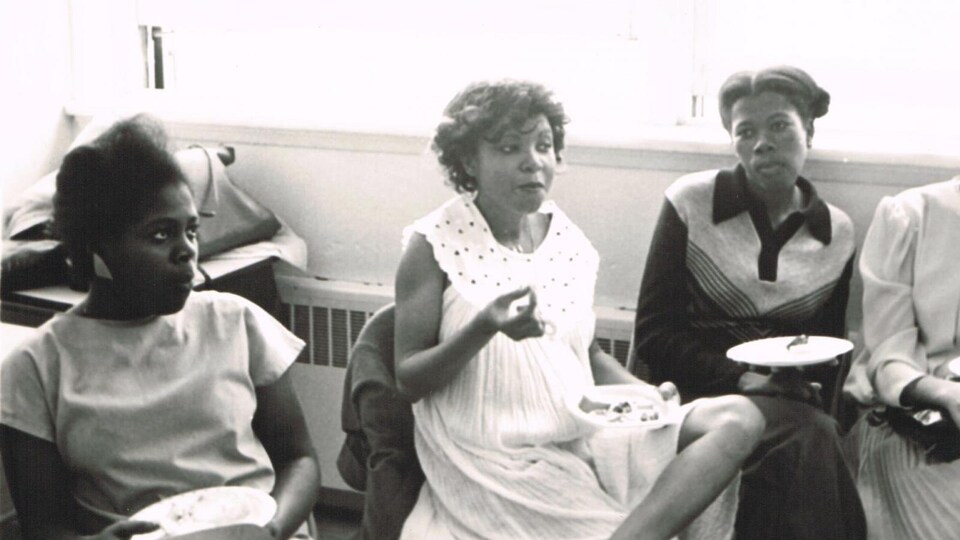 Photo d'archive où on voit des femmes assises dans un local qui discutent en prenant un lunch.