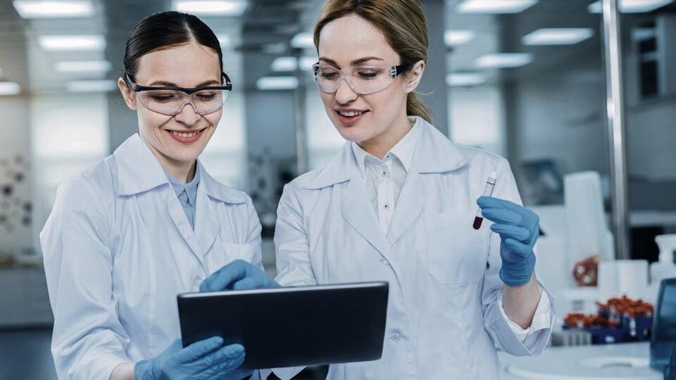 Deux femmes regardent une tablette électronique. Elles portent des gants, des sarraus et des lunettes. Une tient une éprouvette. 
