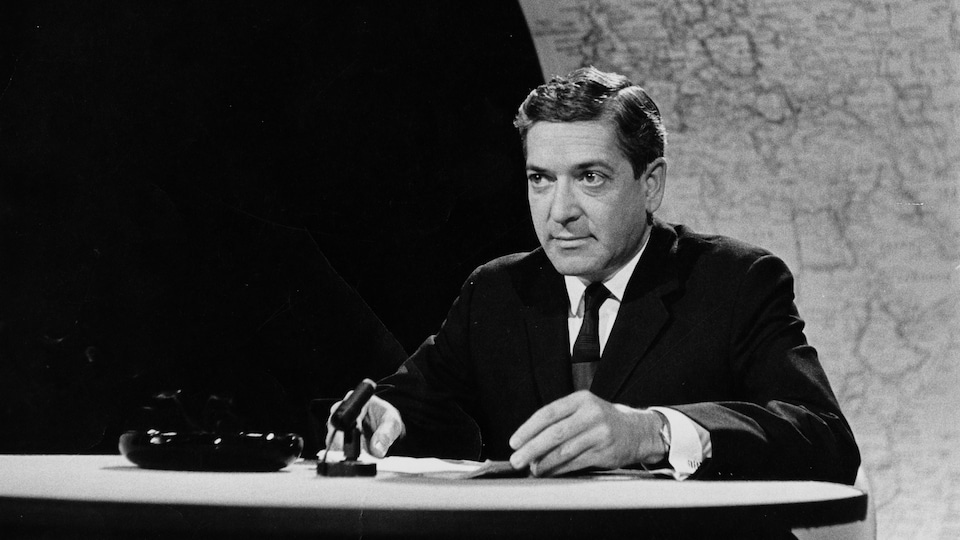 Dans un studio de télévision, Fernand Seguin est assis à un pupitre sur lequel repose un micro de table. En arrière-plan, une immense carte du monde.