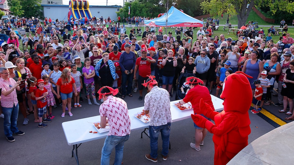 Une foule à l'extérieur autour d'une table où trois personnes décortiquent des homards. Il y a aussi une mascotte de homard.