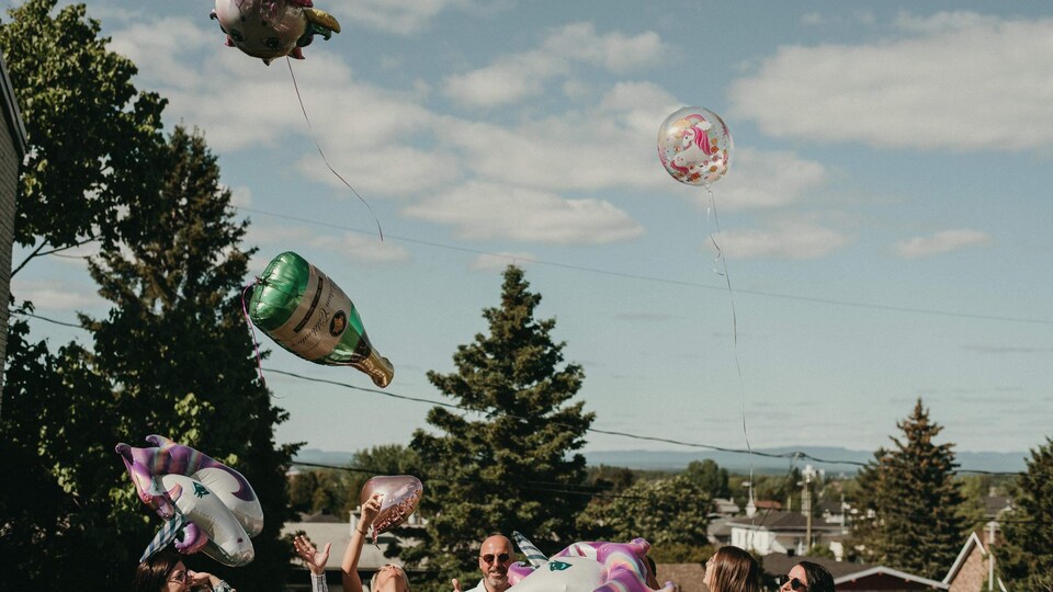 Des ballons à l'hélium dans les airs, en forme de bouteille de champagne et de licorne. On voit le visage de personnes heureuses dans le bas de la photo. C'est l'été, le jour, à l'extérieur.