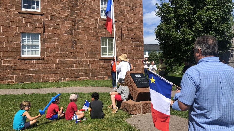 Des personnes s’affichent aux couleurs de l’Acadie et regardent la levée du drapeau acadien devant la banque des fermiers de Rustico, à l’Île-du-Prince-Édouard. 