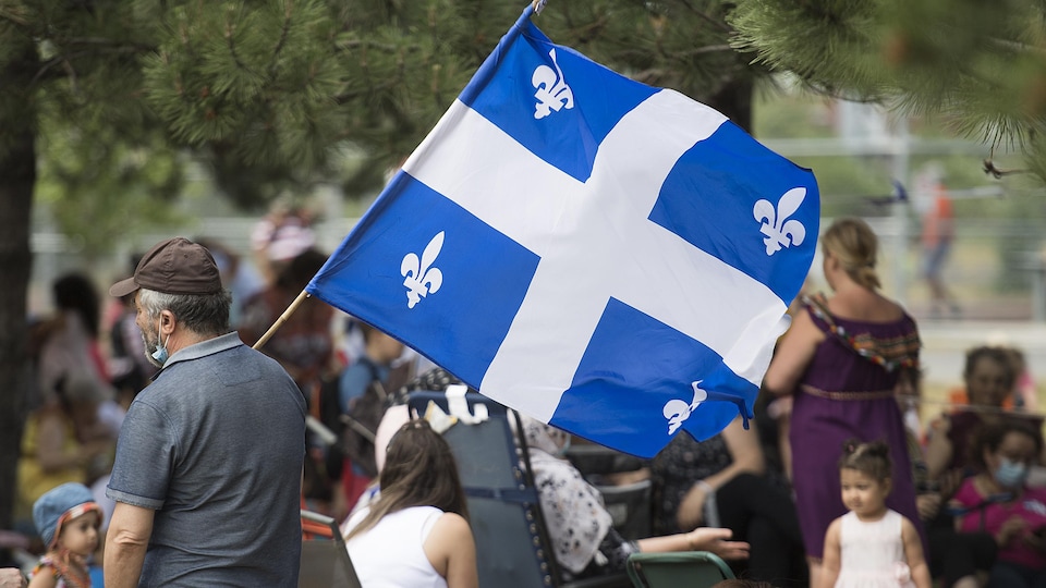 Un homme portant un masque tient un grand drapeau du Québec dans un parc. Derrière lui, des enfants et des femmes sont assis.