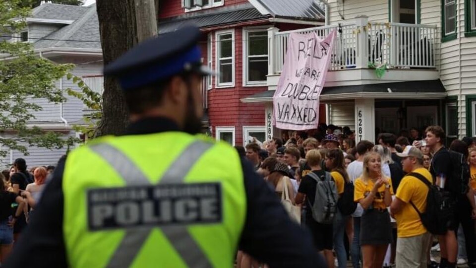 Une police regarde une foule de jeunes près d'une maison dans un quartier résidentiel, où une pancarte affiche « Détendez-vous, nous sommes doublement vaccinés ».