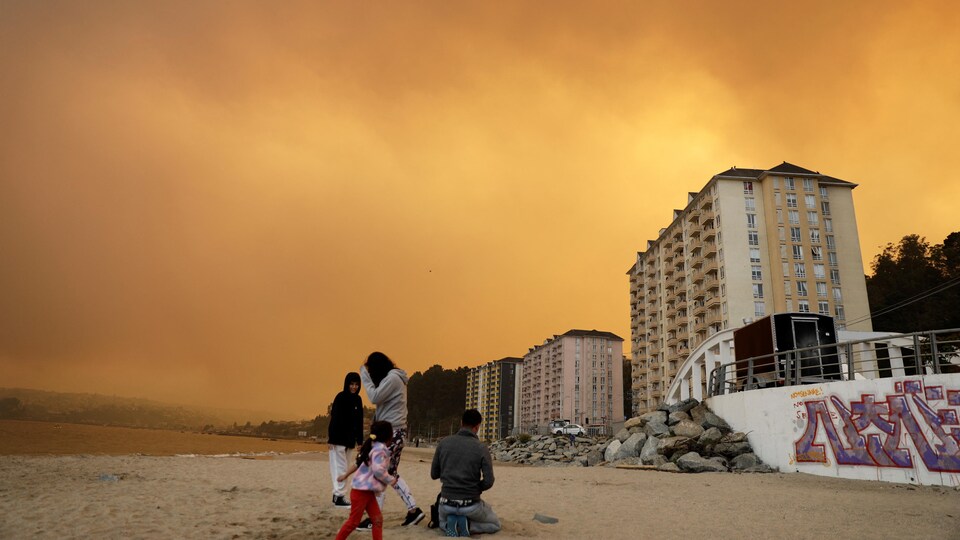 Quatre personnes visitent une plage dans la ville de Tomé au Chili, devant un ciel de brouillard et de fumée.