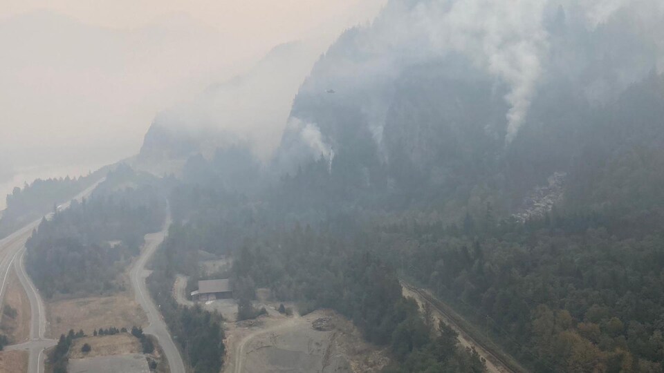Des volutes de fumée s'élèvent d'une forêt située sur le flanc d'une montagne. Le nuage de fumée ainsi formé jette un voile épais sur une route et la voie ferrée qui longe une rivière à la base de la montagne.