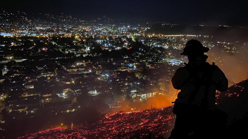 Un pompier, de nuit, devant un feu qui surplombe une vallée très habitée.