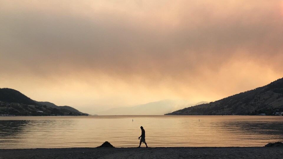 Une silhouette marche près d'un lac sous un ciel enfumé et teinté par les feux de forêt.