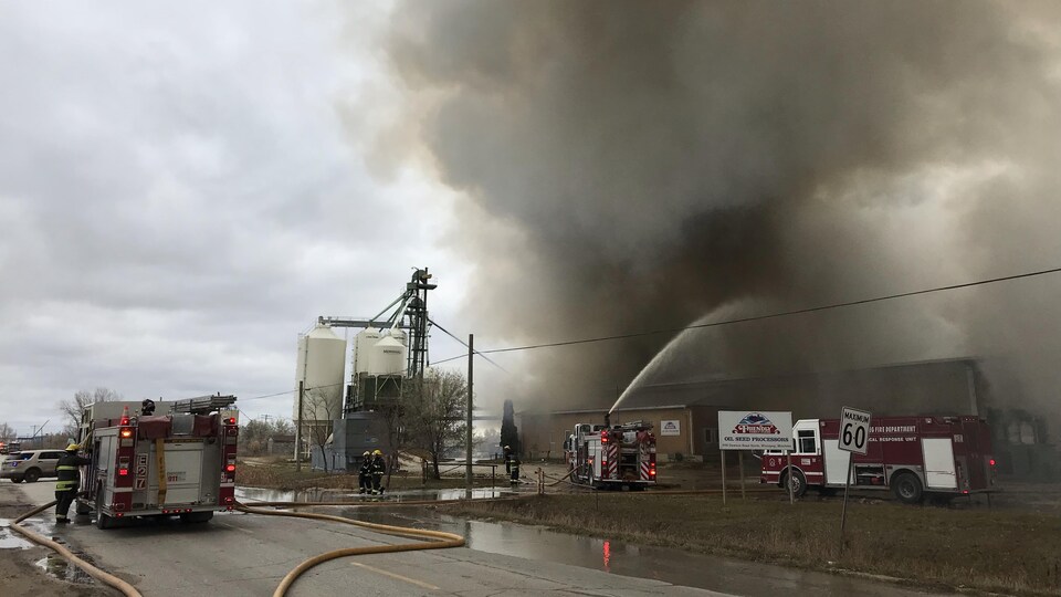 Au bord d'une route se trouvent des silos blancs et un bâtiment duquel s'échappe une épaisse fumée noire. Trois camions de pompiers sont à proximité, l'un d'eux projetant un jet d'eau sur la fumée.