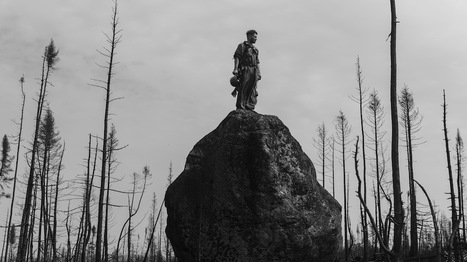 Photo en noir et blanc montrant un homme perché sur le haut d'une grosse roche, entouré d'arbres décimés par le feu. 