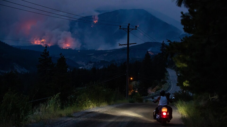 Un homme se promène en motocyclette alors que des montagnes entourant le village de Lytton brûlent.