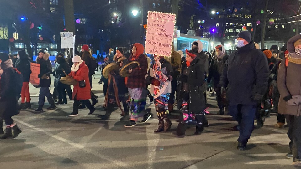 Des personnes, habillées pour affronter le froid, marchent dans une rue. Certaines tiennent des tambours traditionnels et d'autres tiennent des pancartes.