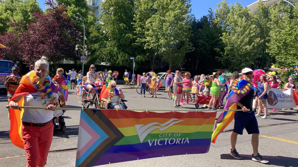 Des personnes vêtues de drapeaux arc-en-ciel tiennent une pancarte de la Ville de Victoria.