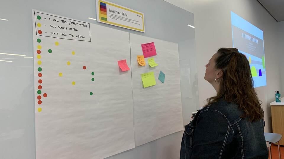 Une femme lit des commentaires écrits sur des bouts de papier sur un tableau blanc, parsemé de collants de diverses couleurs.