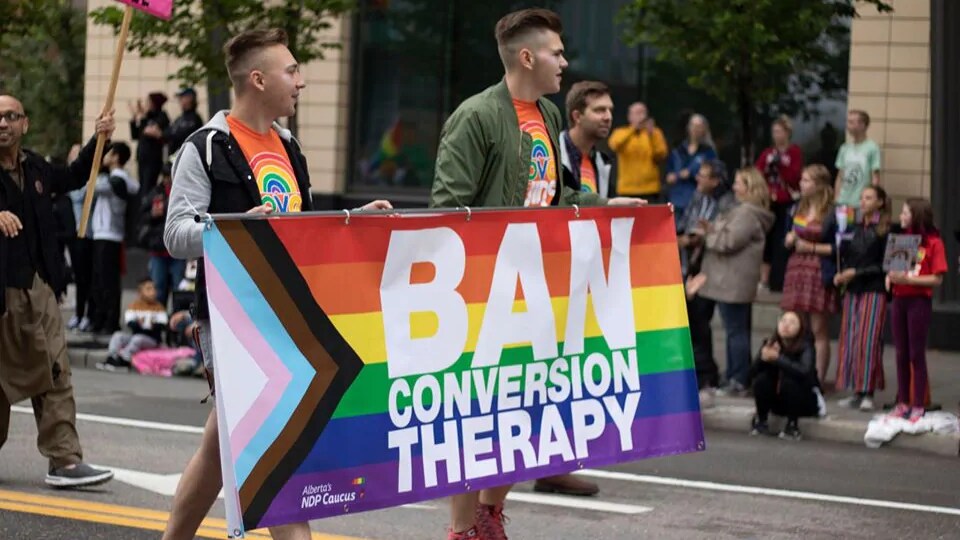 Deux hommes tiennent une bannière où l'on peut lire en anglais : « Interdisez les thérapies de conversion ».