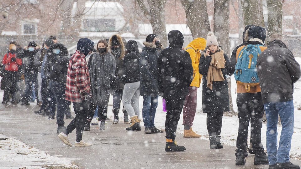 Plusieurs personnes font la file sur un trottoir, sous la neige. 