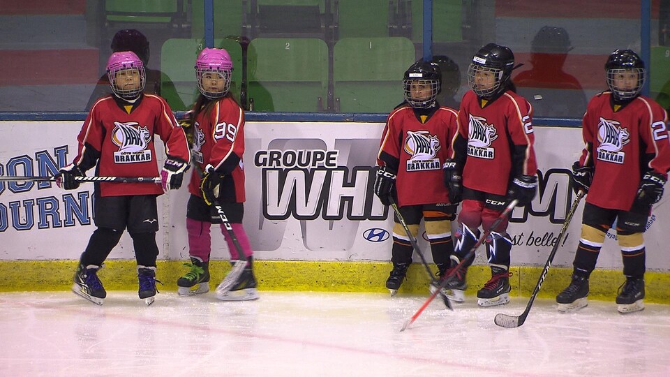 Cinq joueuses de hockey, âgées entre 6 et 8 ans, attendent sur la patinoire.