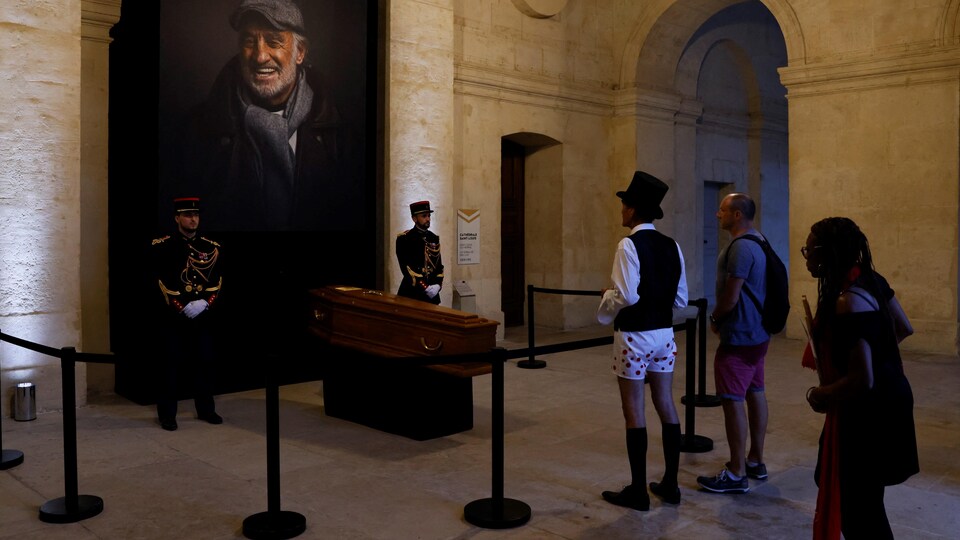 Des personnes sont devant le cercueil de l'acteur dont une photo grand format est en arrière-plan.