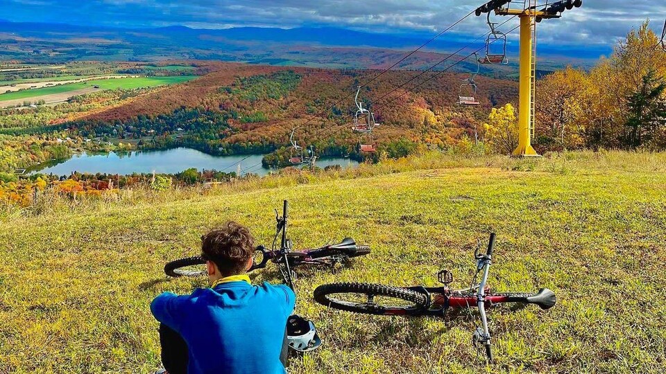 Le fils de Claudie Ouellet vu de dos sur une montagne de ski l'automne. Il est assis près de deux vélos de montagne.