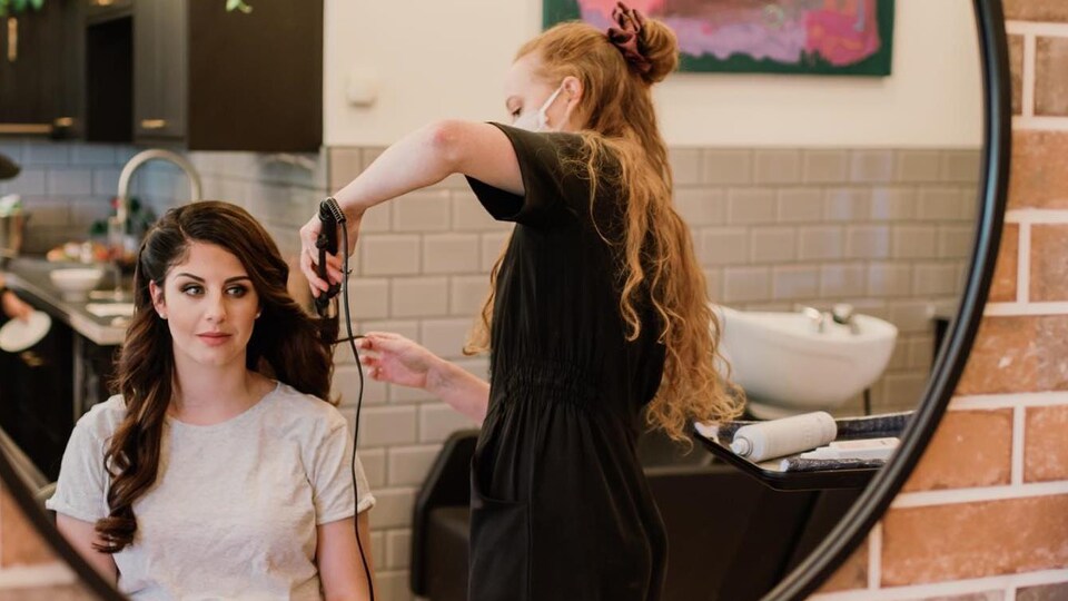 Une coiffeuse qui porte un masque boucle les cheveux d'une jeune femme.