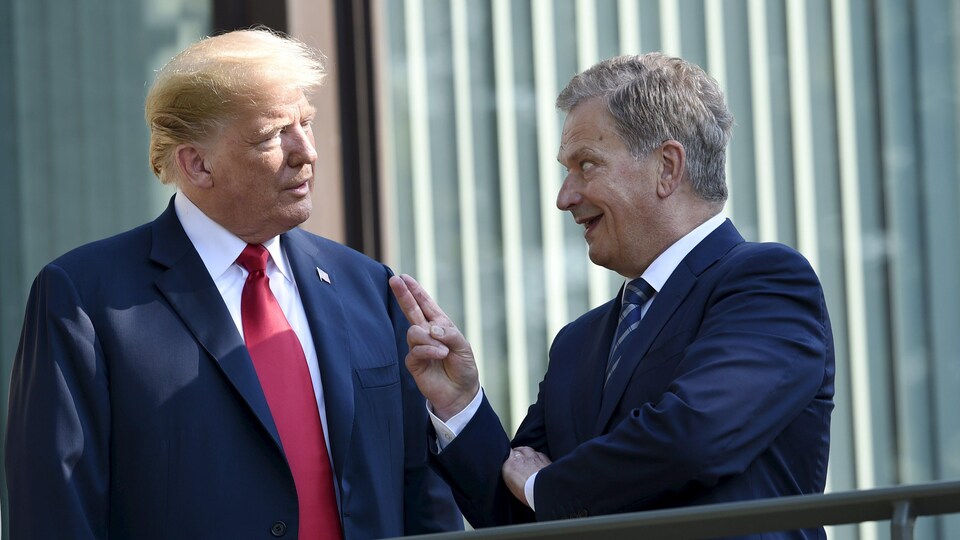 Deux hommes en complet discutent sur le balcon d'un immeuble. L'un d'eux pointe deux doigts vers le ciel en souriant.