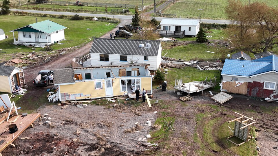 Photo aérienne de chalets et de maisons mobiles endommagées par l'ouragan. Au milieu, une maison n'a plus de toit et deux hommes sont debout devant un grand trou dans le mur.
