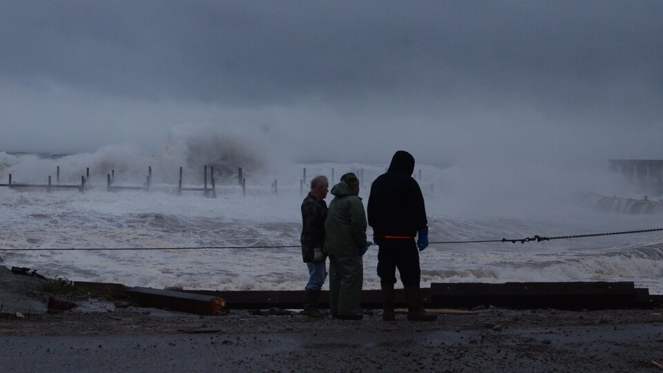 Trois silhouettes sont tournées vers la mer. De très hautes vagues frappent le quai pendant une tempête.